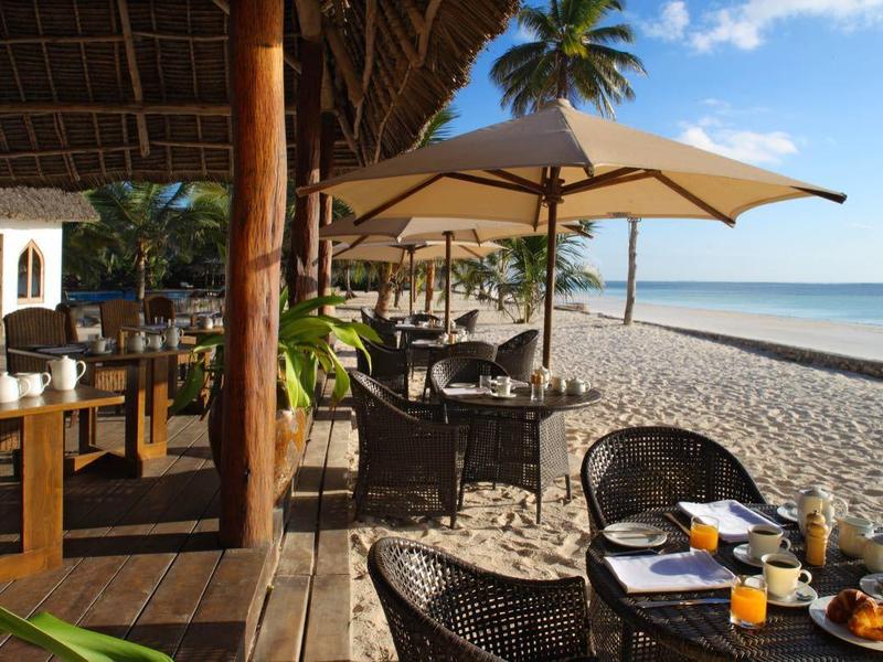 Strandrestaurant mit Tischen, Sonnenschirmen und Meerblick unter blauem Himmel.