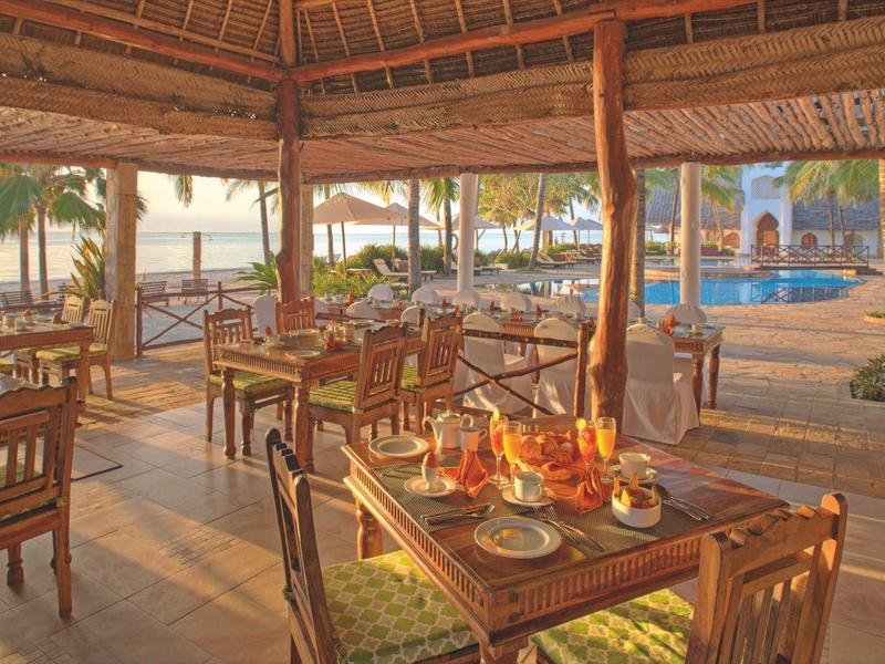 Offener Restaurantbereich mit Holzmöbeln, Blick auf Palmen und Swimmingpool bei Sonnenuntergang.