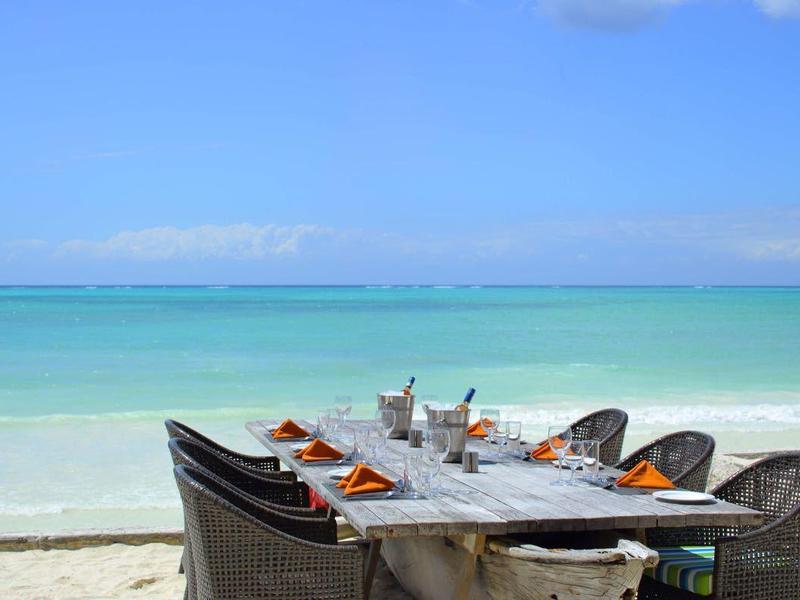 Ein gedeckter Tisch mit Stühlen am Strand mit Blick auf das türkisfarbene Meer.