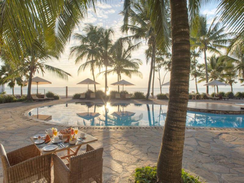 Ruhebereich mit Tisch und Stühlen neben einem Pool mit Palmen und Sonnenschirmen bei Sonnenuntergang.