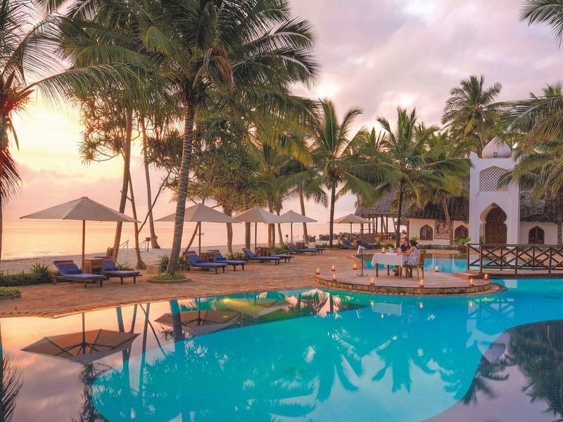Luxuriöser Poolbereich mit Palmen und Sonnenliegen am Strand bei Sonnenuntergang.
