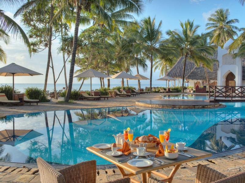 Tropischer Poolbereich mit Tischen, Stühlen und Palmensoase am Strand bei Sonnenuntergang.