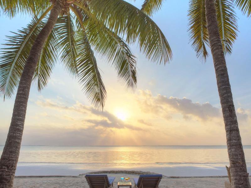 Zwei Liegestühle mit einem Tisch dazwischen am Strand unter Palmen bei Sonnenuntergang.