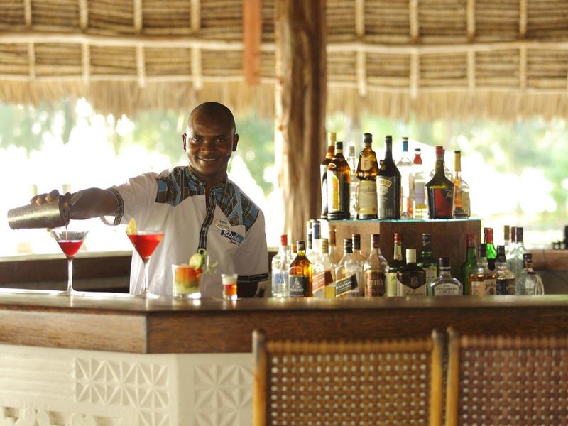 Barkeeper gießt Getränke in zwei Cocktails an einer offenen Bar mit Flaschen im Hintergrund.