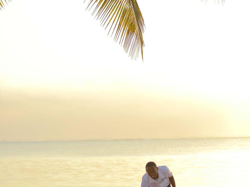 Zwei Personen neben zwei Liegestühlen am Strand unter Palmenzweigen bei Sonnenuntergang.