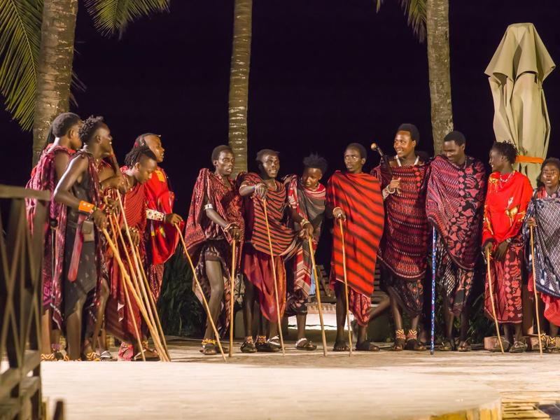 Gruppe von Menschen in traditioneller Kleidung steht nachts unter Palmen auf einem Holzsteg.