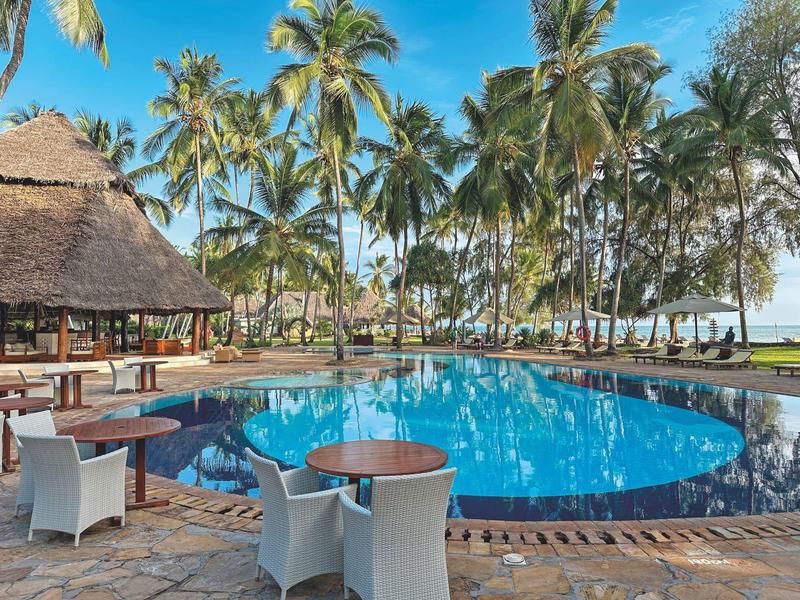 Runder Pool mit Sitzbereichen unter Palmen am Strand bei klarem Himmel.