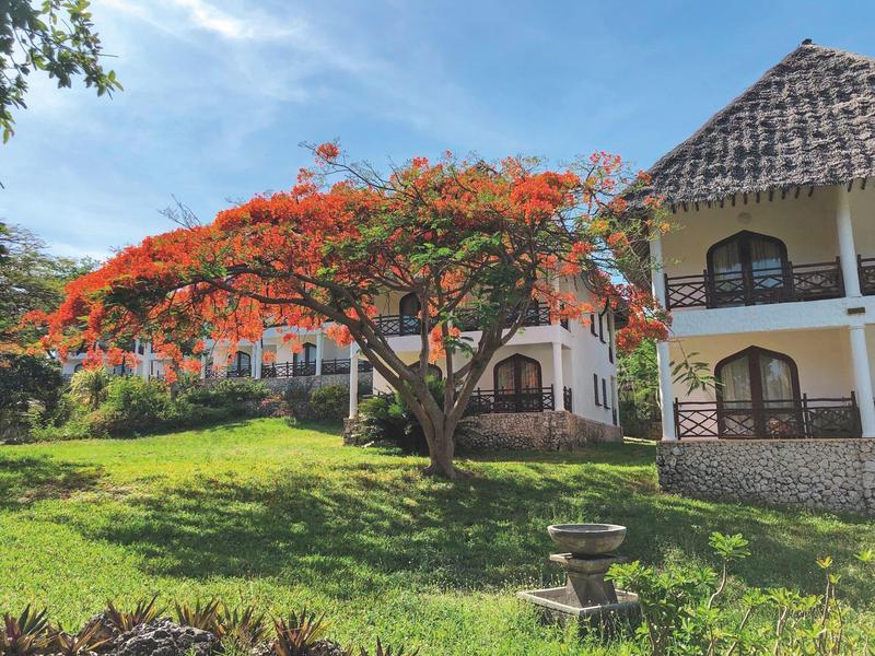 Haus mit steinernem Dach neben einem Baum mit roten Blüten und grünem Rasen.