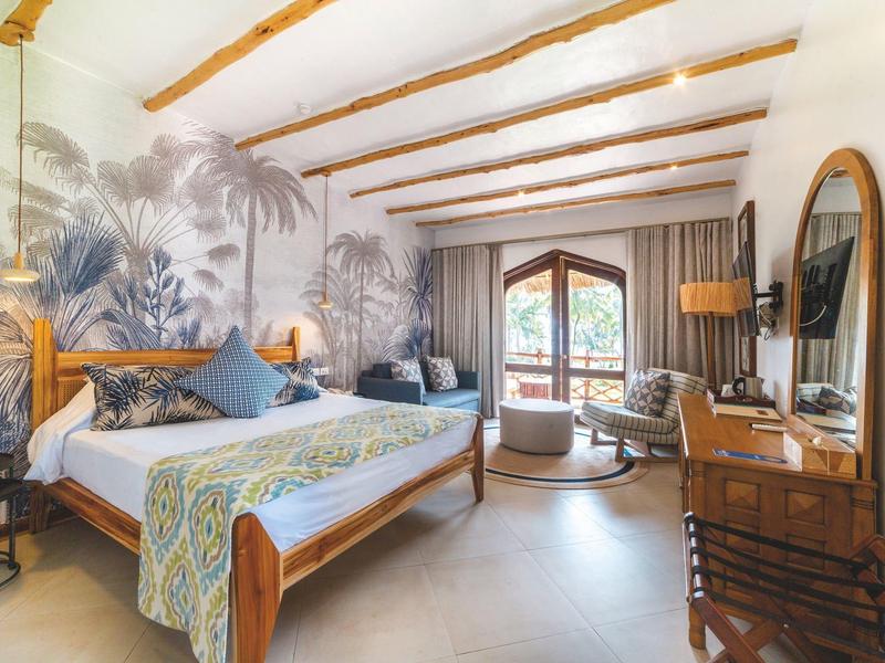 Hell eingerichtetes Hotelzimmer mit Holzbalken, großem Bett und tropischer Wandgestaltung.
