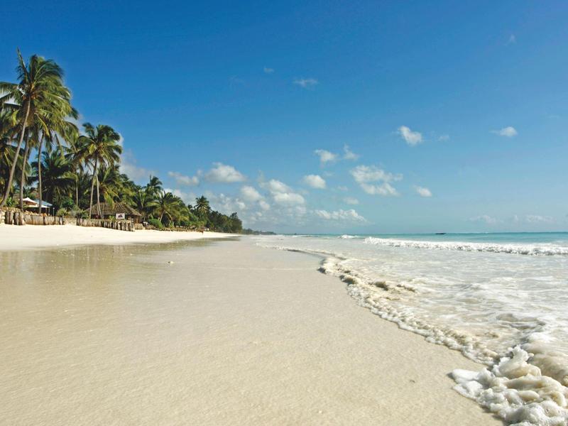 Breiter Sandstrand mit Palmen, blauem Himmel und sanften Wellen am Meer.