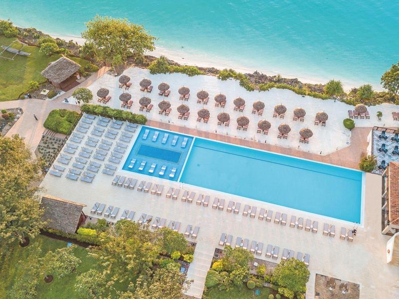 Großer rechteckiger Pool mit blauen Liegen und Sonnenschirmen neben türkisfarbenem Wasser und Bäumen.