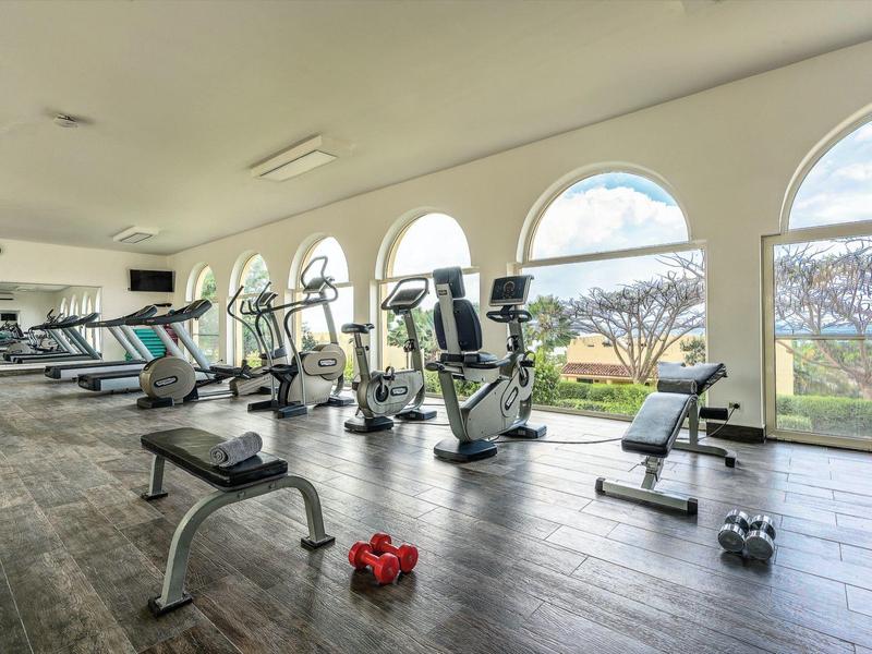 Hell erleuchteter Fitnessraum mit großen Rundfenstern, Trainingsgeräten und Hanteln auf Holzfußboden.