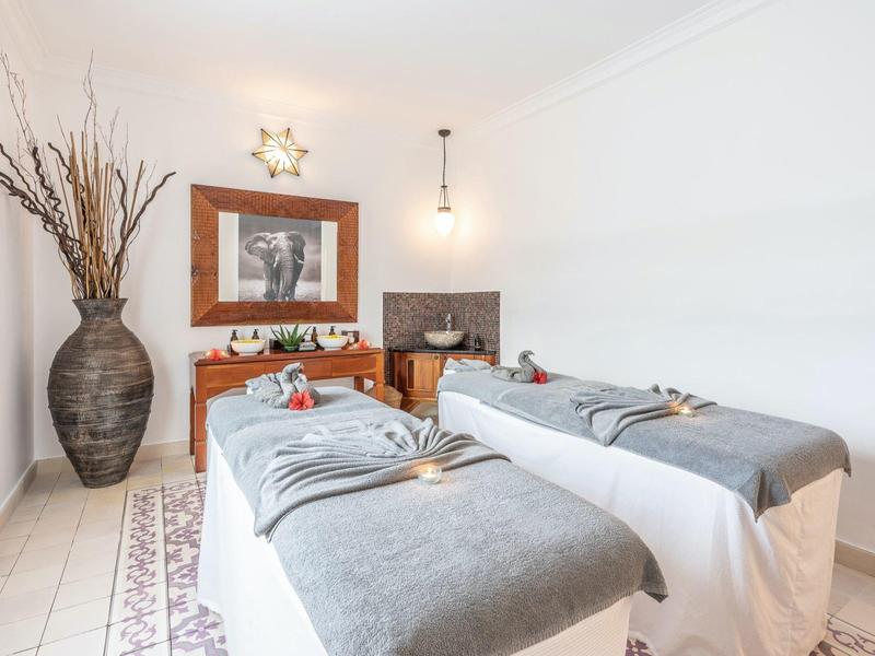 Zwei Massageliegen mit grauen Decken in hellem, modernem Raum mit Vase, Spiegel und Holzkommode.