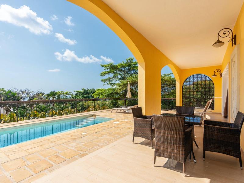 Terrasse mit gelben Wänden, runden Bögen, braunen Stühlen und Pool unter blauem Himmel.