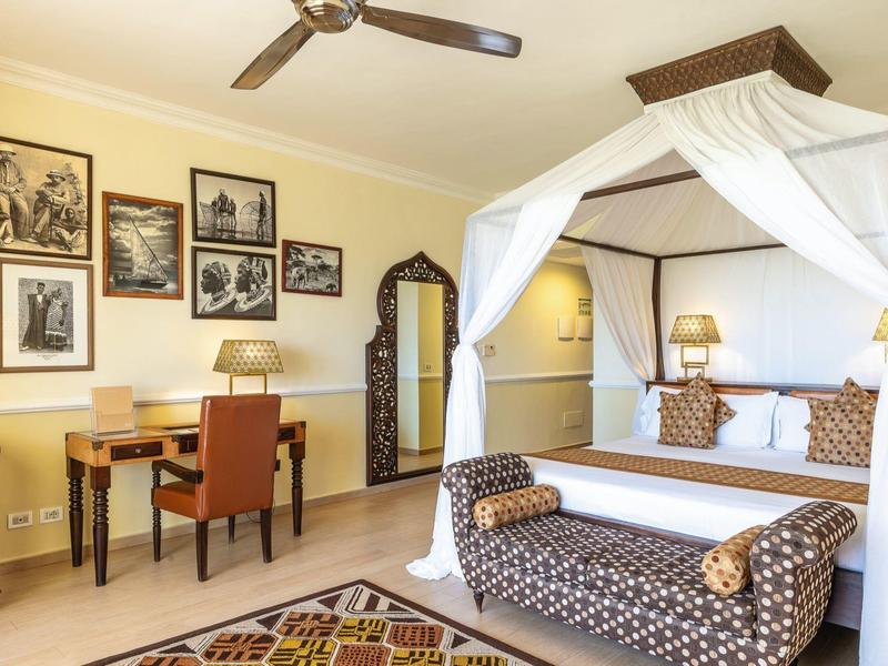 Hell eingerichtetes Schlafzimmer mit himmelbett, gemütlicher Sitzbank und Schreibtisch am Fenster.