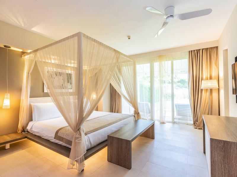 Helles Schlafzimmer mit Himmelbett, Holzbank, großen Fenstern und beigem Vorhang.