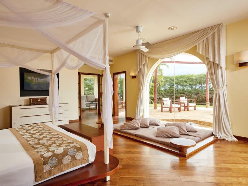 Helles Schlafzimmer mit Himmelbett, großem Fenstertor und Sitzkissen auf Holzboden.