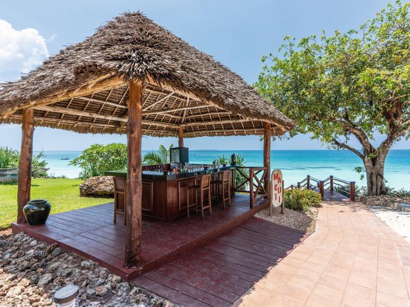 Offener Holzpavillon mit Strohdach und Bar, davor Baum, Strand und türkisfarbenes Meer.