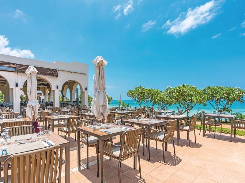 Große Terrasse mit Holztischen und Stühlen, blauer Himmel, Meerblick und Sonnenschirmen.