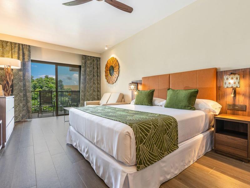 Modernes Hotelzimmer mit großem Bett, grünen Kissen, Balkon und Deckenventilator.