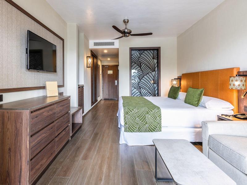 Helles, modernes Schlafzimmer mit Doppelbett, Holzmöbeln, Deckenventilator und Sitzbereich am Fenster.