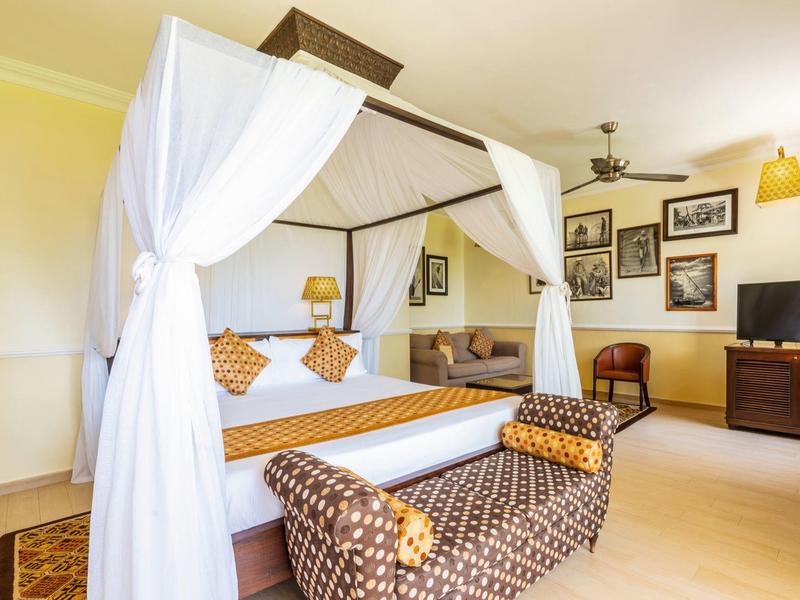 Helles Schlafzimmer mit Himmelbett, gemustertem Sofa, Fernseher und Wandbildern in warmen Tönen.