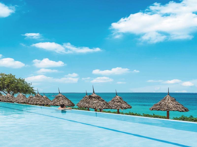 Infinity-Pool mit blauem Wasser, dahinter grüne Palmen und türkisfarbenes Meer unter blauem Himmel.