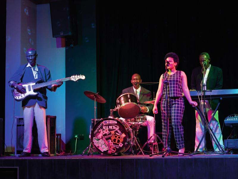 Vierköpfige Band mit Gitarrist, Schlagzeuger, Sängerin und Keyboarder auf dunkler Bühne.