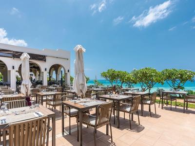 Hotelterrasse mit Holztischen, Sonnenschirmen und Blick auf das blaue Meer bei Sonnenschein.