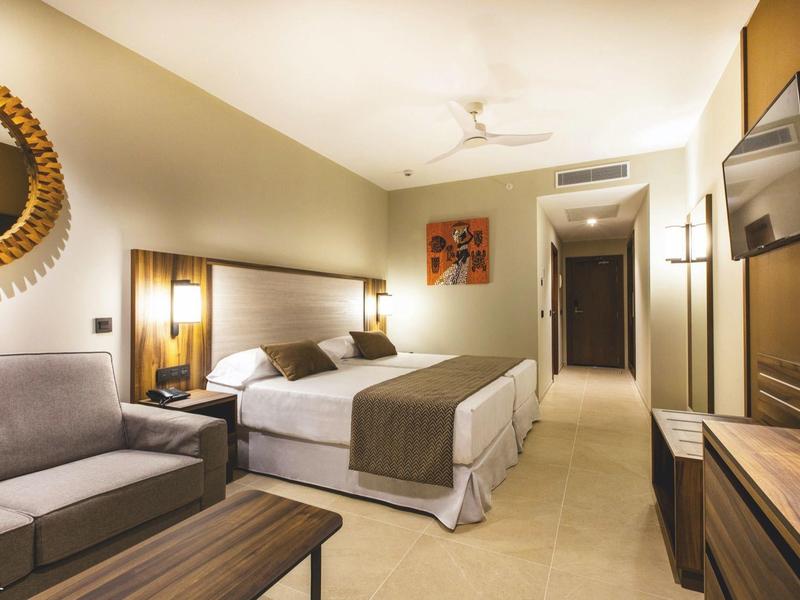 Moderner Hotelzimmer mit Doppelbett, Sofa, Spiegel, TV und warmem Licht im hellen Raum.