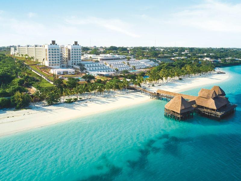 Strand mit klarem türkisfarbenem Wasser, weißen Sand und Gebäuden am Ufer unter blauem Himmel.