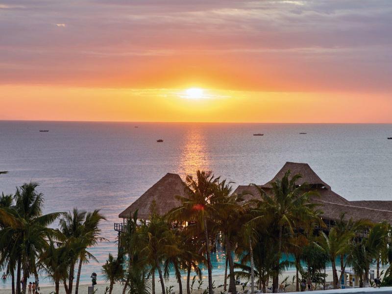 Sonnenuntergang über dem Meer mit Hütten und Palmen im Vordergrund, sanfte Wolken am Himmel.