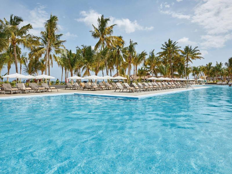 Großer Pool mit klar blauem Wasser, umgeben von Palmen und weißen Liegestühlen unter blauem Himmel.