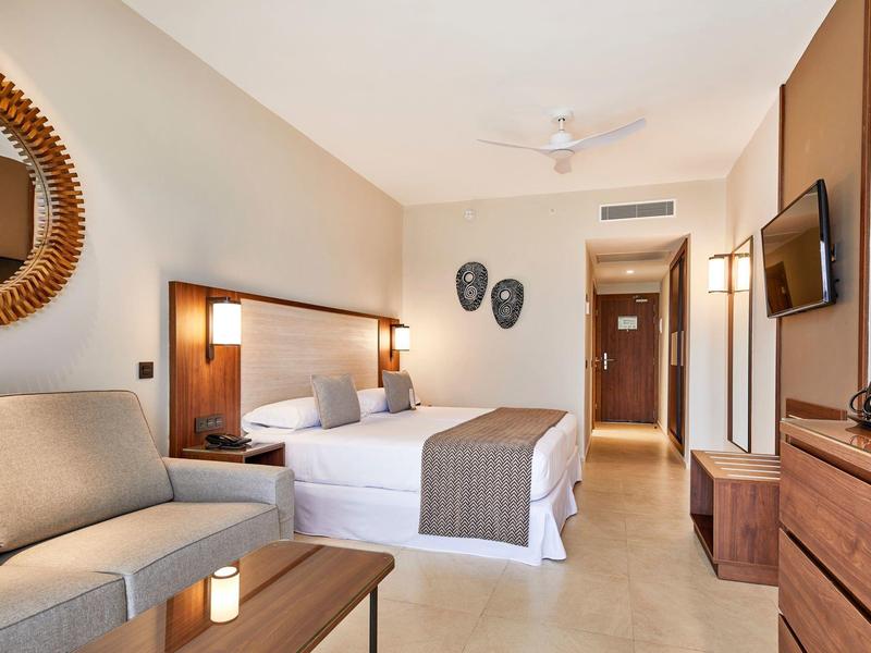 Helles Hotelzimmer mit Doppelbett, Sofa, Wandspiegel, TV und Holzmöbeln in warmen Farbtönen.