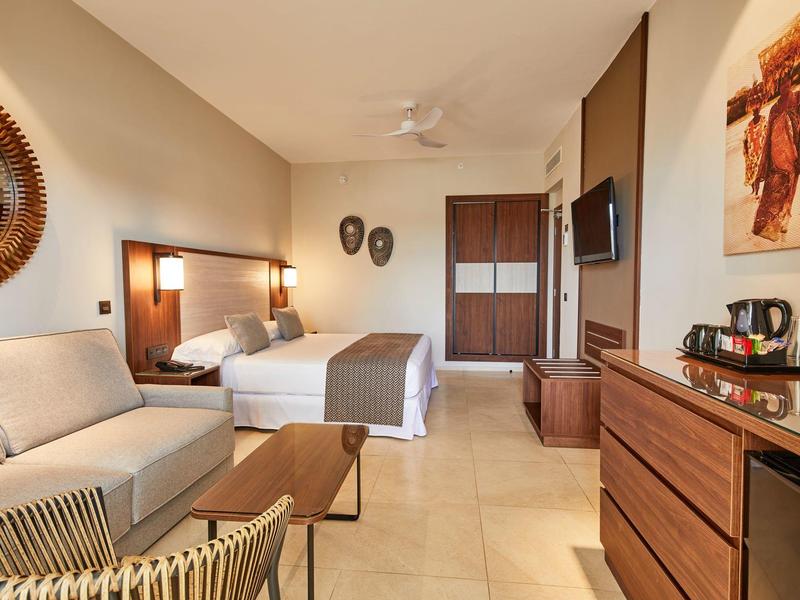 Modern eingerichtetes Hotelzimmer mit Doppelbett, Sofa, Schrank und warmen Holzmöbeln.