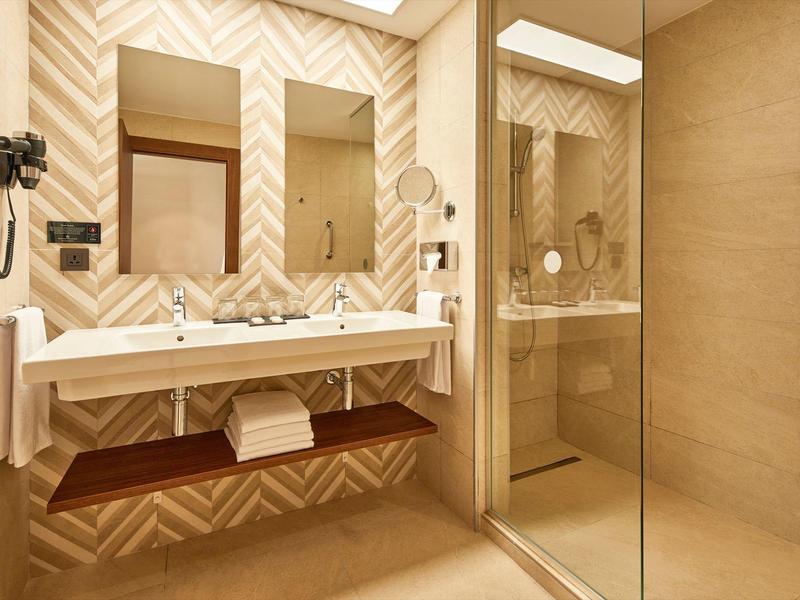 Modernes Badezimmer mit Doppelwaschbecken, Glasscheibe zur Dusche und hellen Kacheln in Beige.