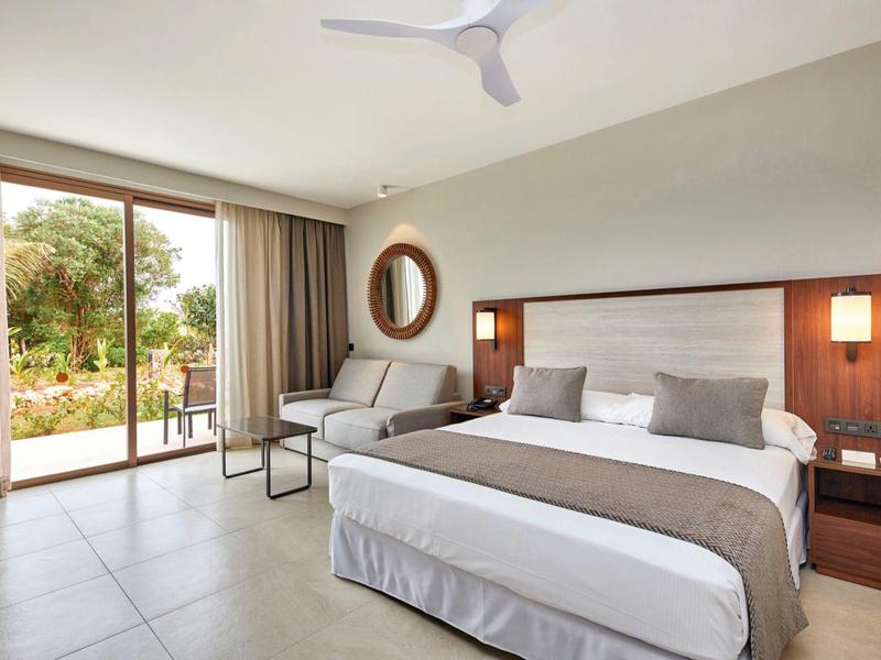 Helles Schlafzimmer mit großem Bett, hellgrauem Sofa, Glas-Schiebetür und Blick ins Grüne.