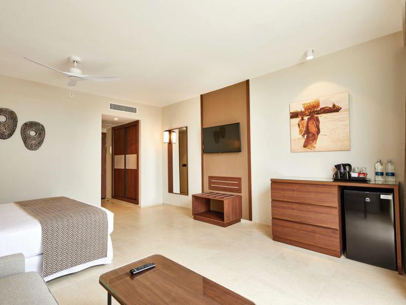 Helles Hotelzimmer mit Doppelbett, Holzmöbeln, TV, Minibar und moderner Wanddekoration.