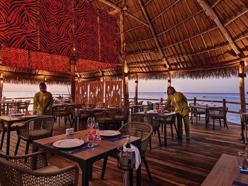 Offenes Restaurant mit Holzdach, Stühlen, Tischen und Meerblick bei Sonnenuntergang.