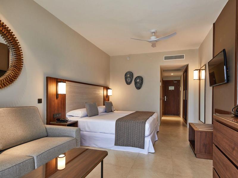 Modernes Hotelzimmer mit Doppelbett, Sofa, Wandspiegel, Deckenventilator und Flachbildfernseher.