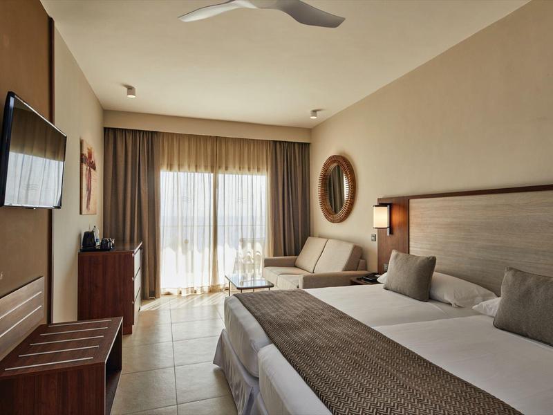 Helles Hotelzimmer mit Doppelbett, Fernseher, Holzmöbeln und Balkon mit Vorhängen.