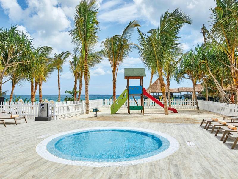 Pool mit rundem Whirlpool, Liegestühlen, Palmen und Kinderspielplatz am Meer unter blauem Himmel.