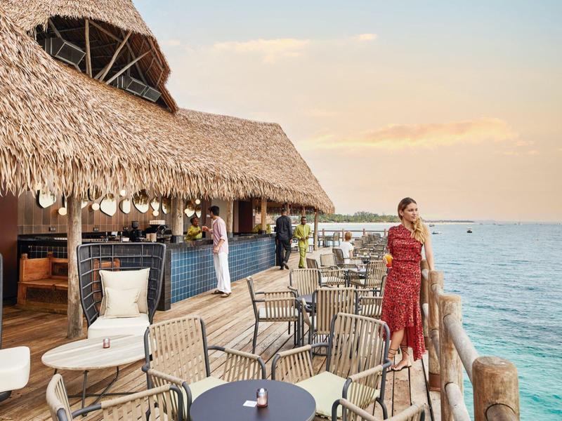 Terrassenrestaurant am Meer mit Stroh gedecktem Dach, Tischen, Stühlen und zwei Personen bei Sonnenuntergang.