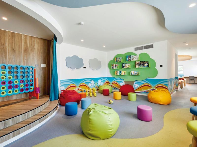 Bunte Kinderspielecke mit Kissen, Hockern und Regalen vor Wand mit Baum- und Wolkenmotiven.