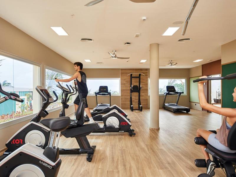 Fitnessraum mit modernen Geräten, hellen Fenstern, Holzfußboden und zwei Personen beim Training.