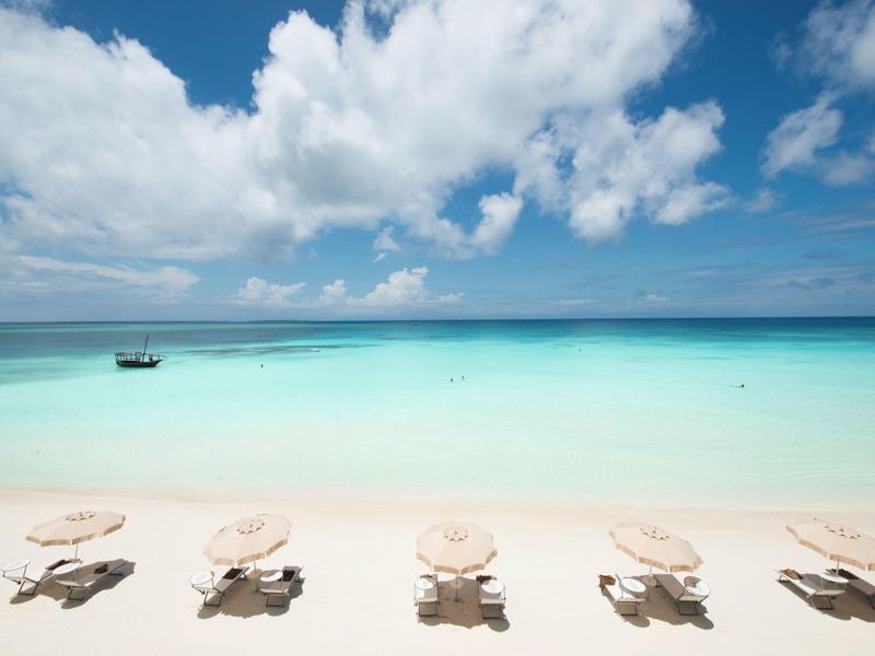 Fünf weiße Strandliegen mit Schirmen stehen auf hellem Sand vor türkis-blauem Meer und blauem Himmel.