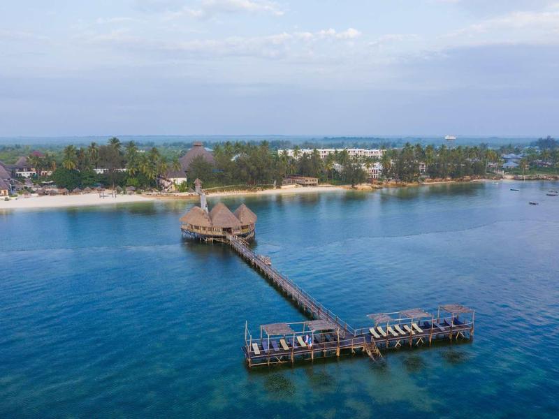 Holzsteg mit Hütten über blauem Meer, im Hintergrund grüner Küstenstreifen und Himmel mit Wolken.