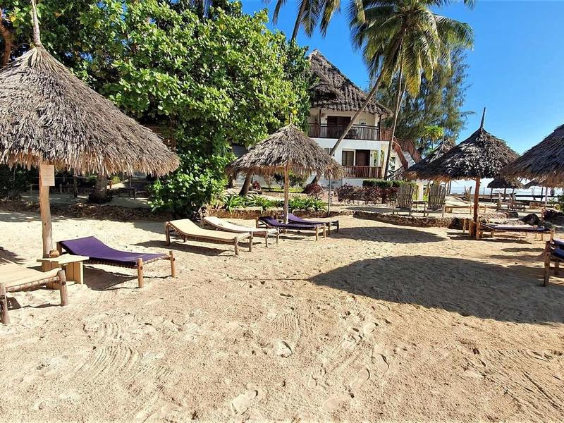 Ein sonniger Strand mit Sand, Holzhütten, Palmen und Liegestühlen unter Strohdächern.