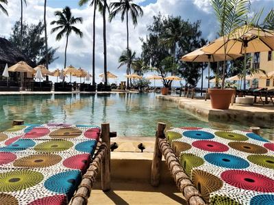 Großer Pool mit bunten Sitzkissen, Palmen, Sonnenschirmen und blauem Himmel im Hintergrund.