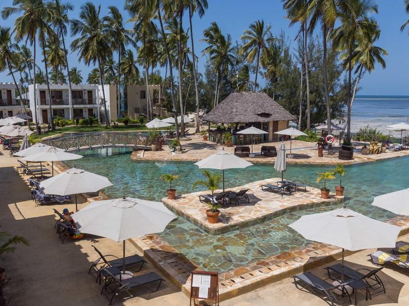 Luxuriöser Hotelpool mit Palmen, Sonnenliegen und Sonnenschirmen am Strand unter blauem Himmel.